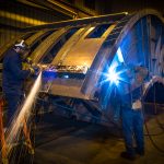 Welding operator welding on large piece of vibrating equipment with another person grinding